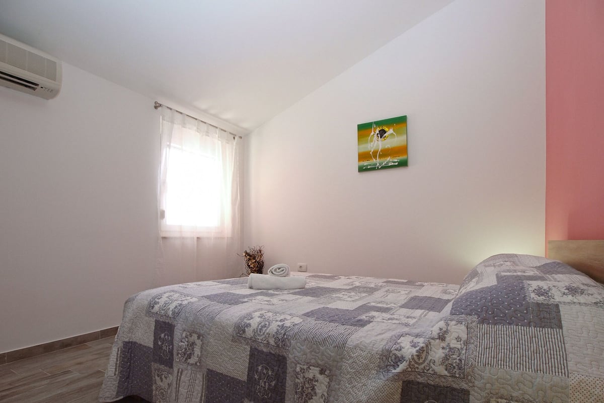 Ein Schlafzimmer mit einem Bett, einem Fenster mit durchsichtigen Vorhängen, einer Klimaanlage und einem Gemälde an der Wand.