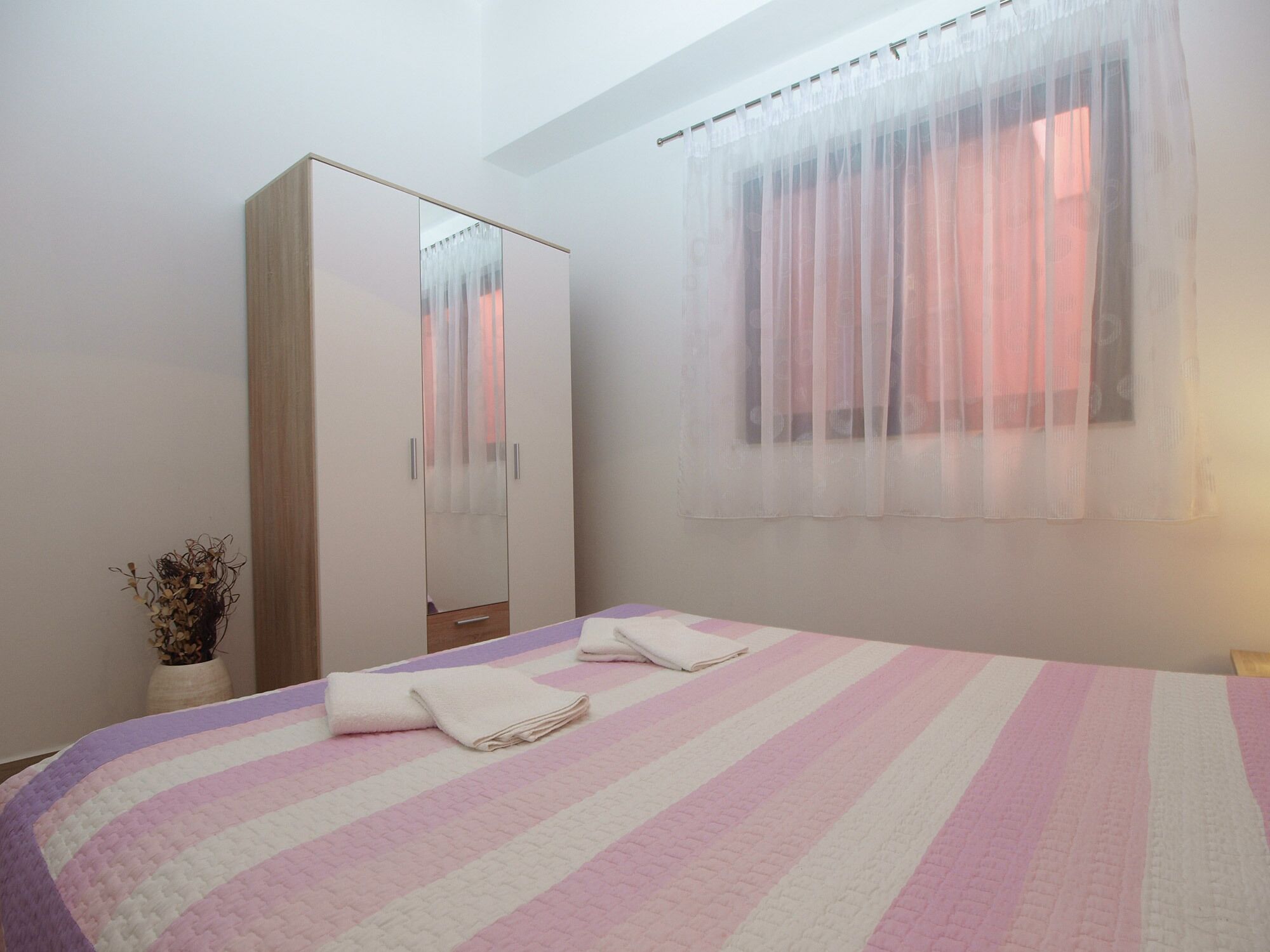A bedroom with a bed, a wardrobe, a window with sheer curtains, and a vase with dried flowers.