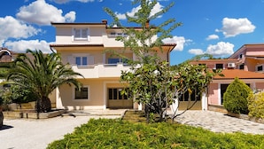 A two-story house with a tiled roof, a front garden with various plants, and a clear blue sky. - Apartments Emil 596 (Pula)