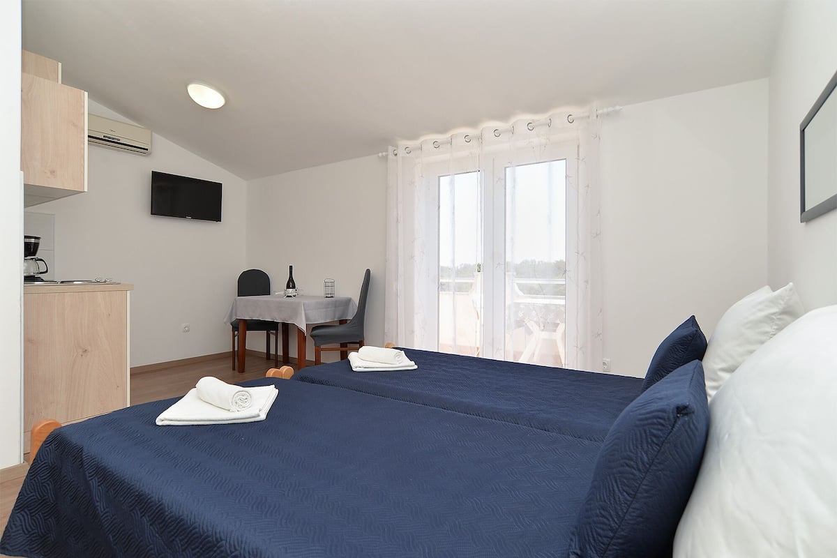 A hotel room with a bed, a desk with a chair, a television mounted on the wall, and a balcony with curtains.