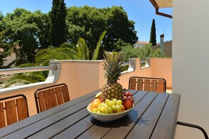 Ein Tisch mit Obstschale, darunter Ananas, Trauben und Äpfel, auf einer Terrassenanlage mit Stühlen und Geländer.