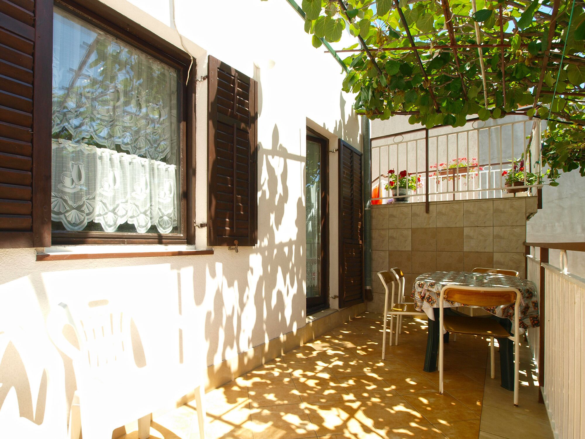 A narrow outdoor corridor with a tiled floor, a table with chairs, and a window with lace curtains.