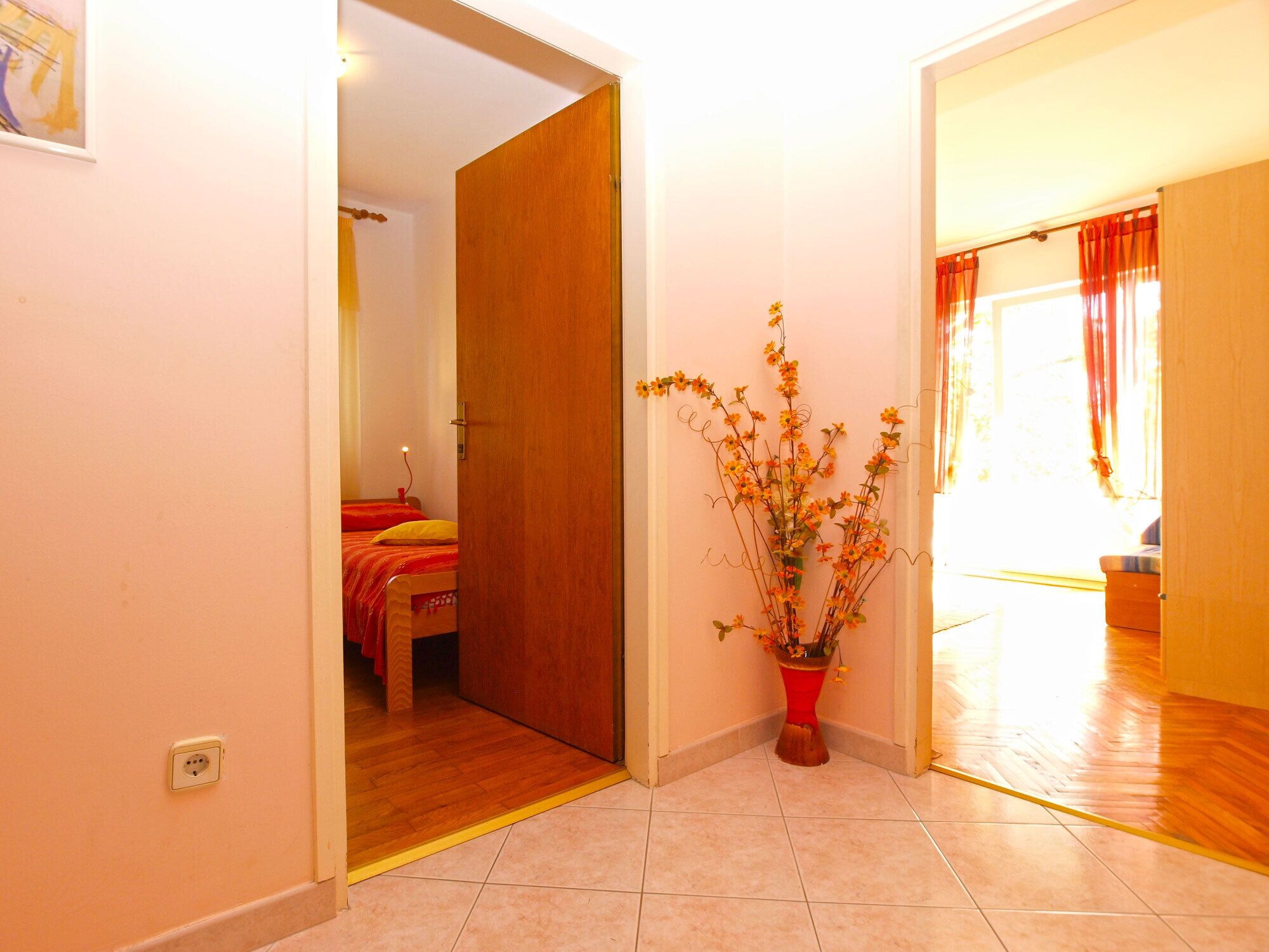 A room with a wooden door, a bed with red bedding, and a potted plant with yellow flowers.