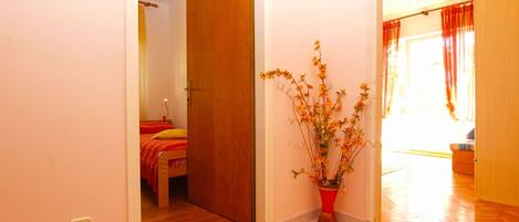 A room with a wooden door, a bed with red bedding, and a potted plant with yellow flowers.
