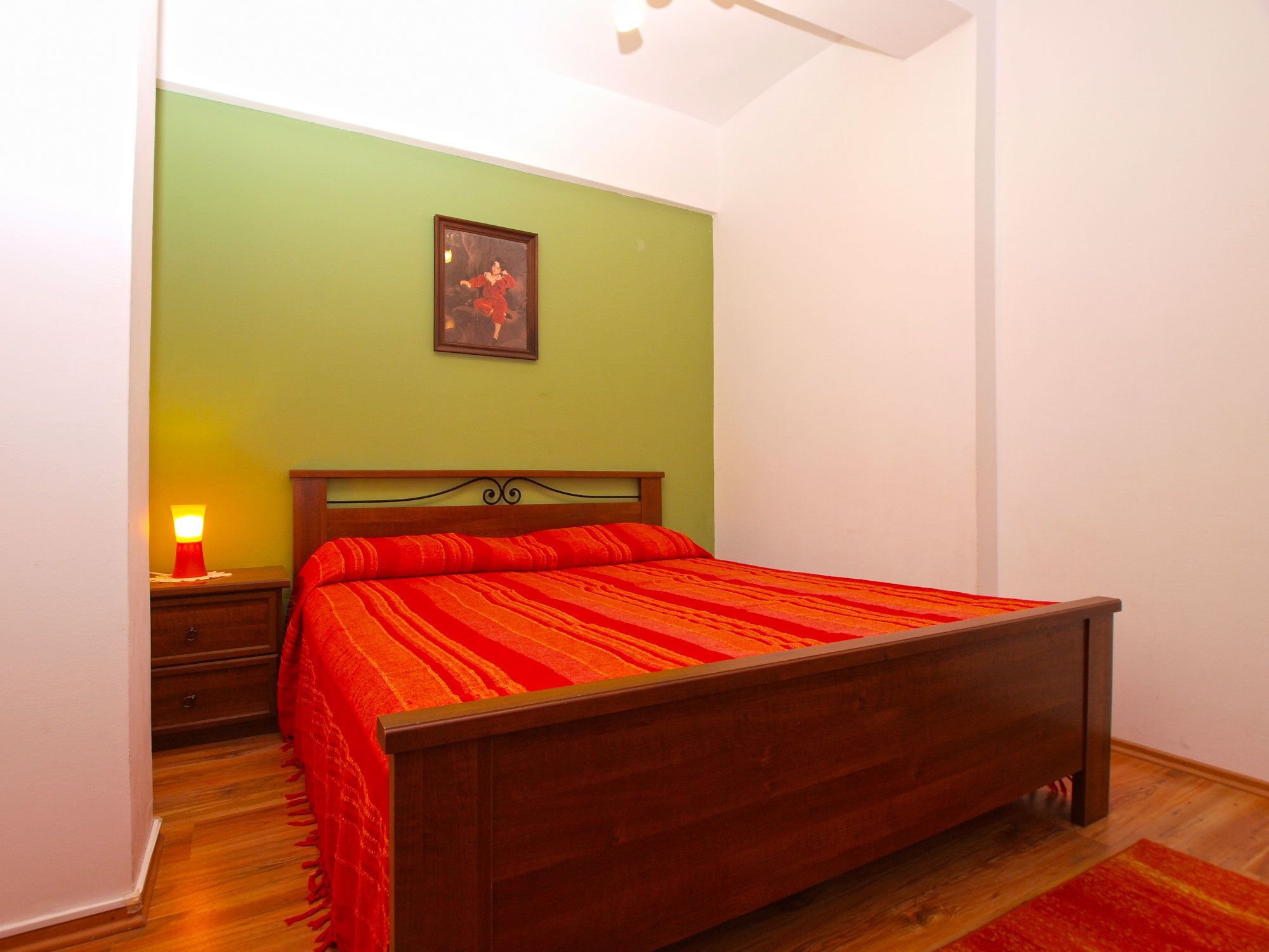Een slaapkamer met een houten bed, nachtkastje, lamp en een lijst met een afbeelding aan de muur.