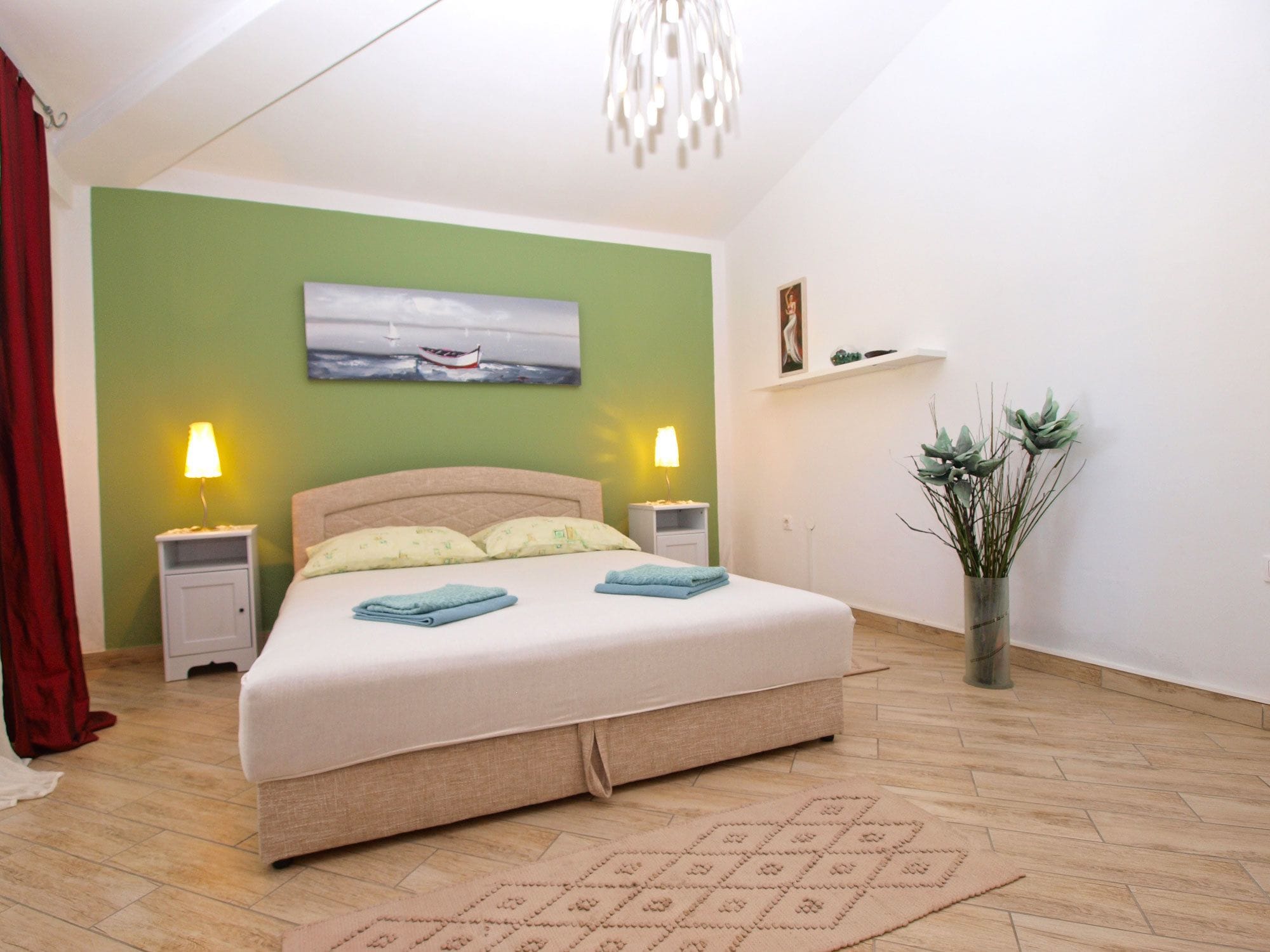 A bedroom with a bed, two bedside lamps, a painting, a plant, and a red curtain.