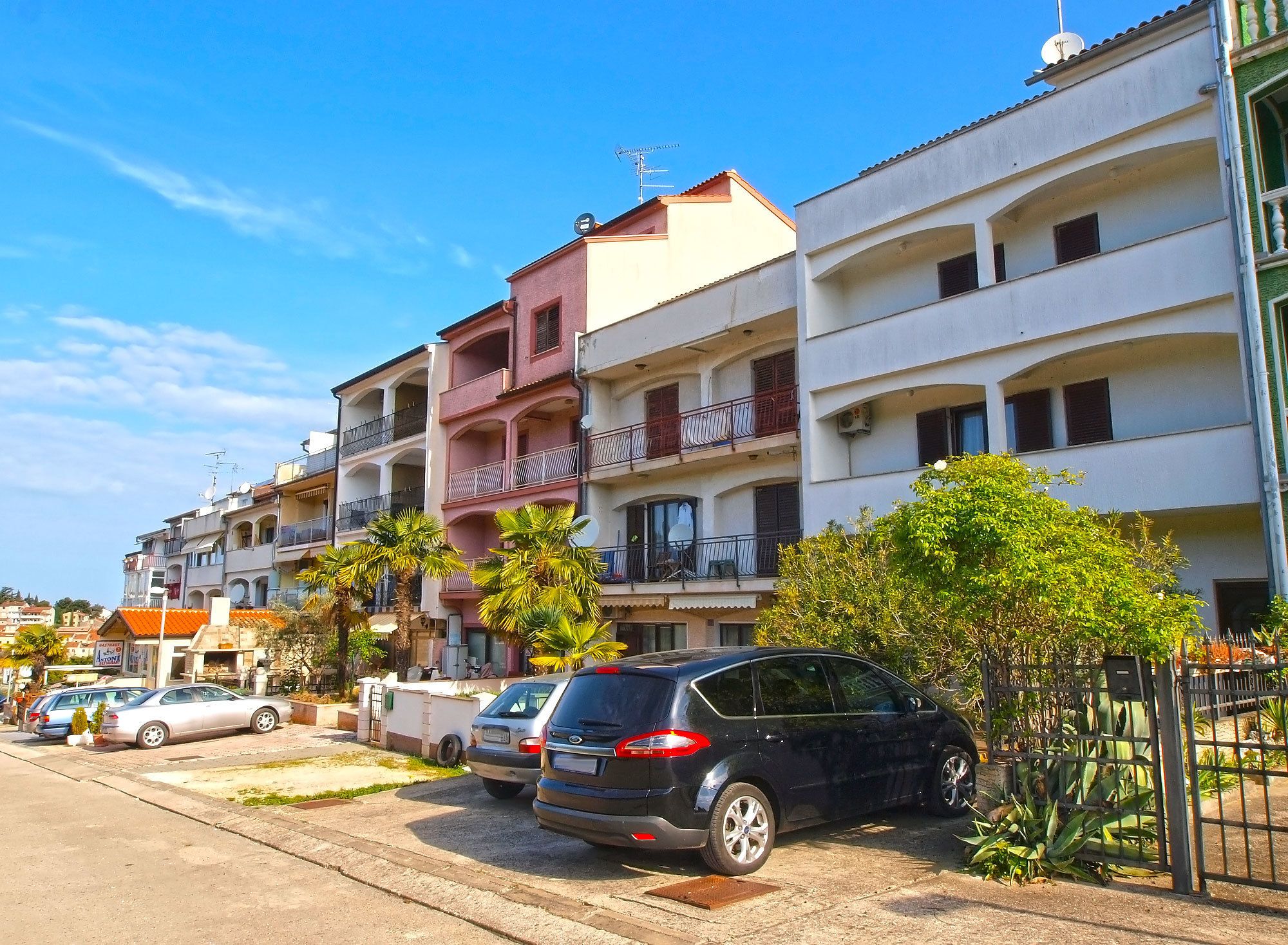 A row of multi-story residential buildings with balconies, parked cars, and a clear blue sky.