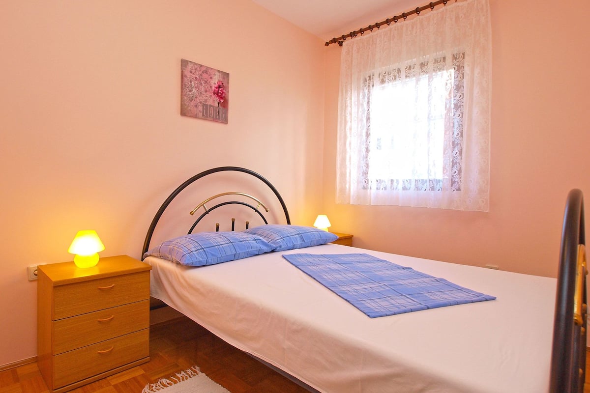 Une chambre à coucher avec un lit, deux lampes de chevet, une table de nuit en bois, un petit tiroir, une fenêtre avec des rideaux en dentelle et un tableau accroché au mur.