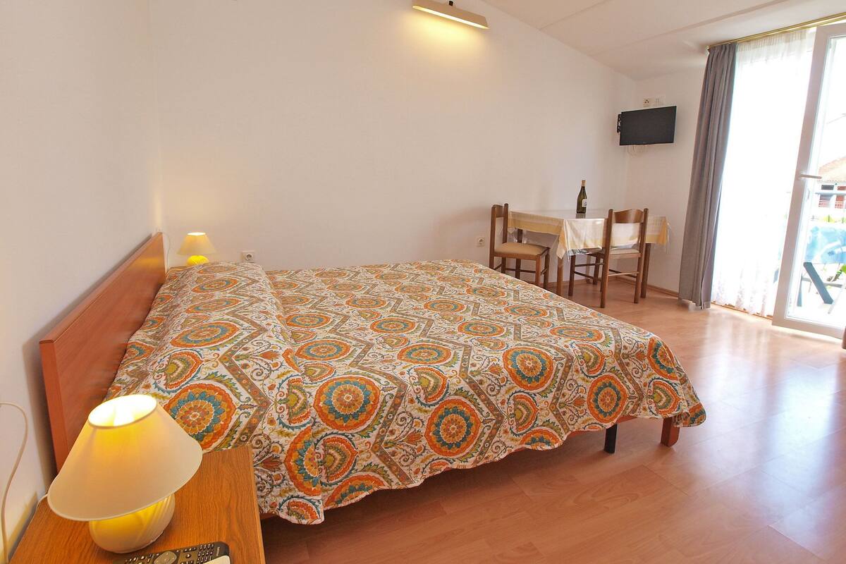 Quarto com cama, mesa de cabeceira com abajur, escrivaninha com cadeira e televisão na parede.