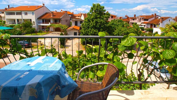 Een balkon met een rieten stoel en een tafel bedekt met een blauwe tafelkleed, met uitzicht op een woongebied met huizen en groen.