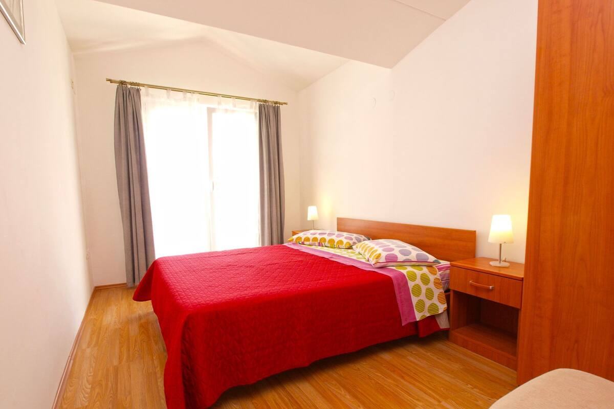 Um quarto bem arrumado com colcha vermelha, mesa de cabeceira de madeira com abajur e janela com cortinas.
