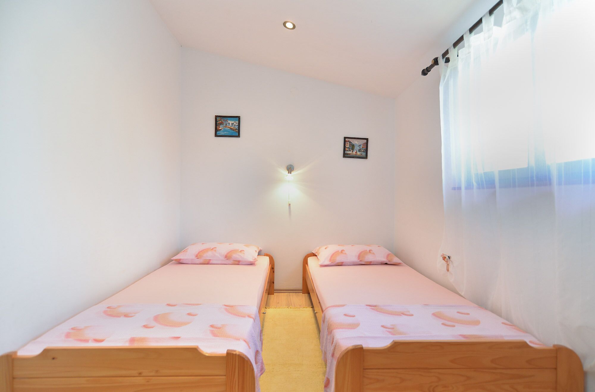 A small, clean room with two wooden beds, a wall-mounted light, and two small pictures.