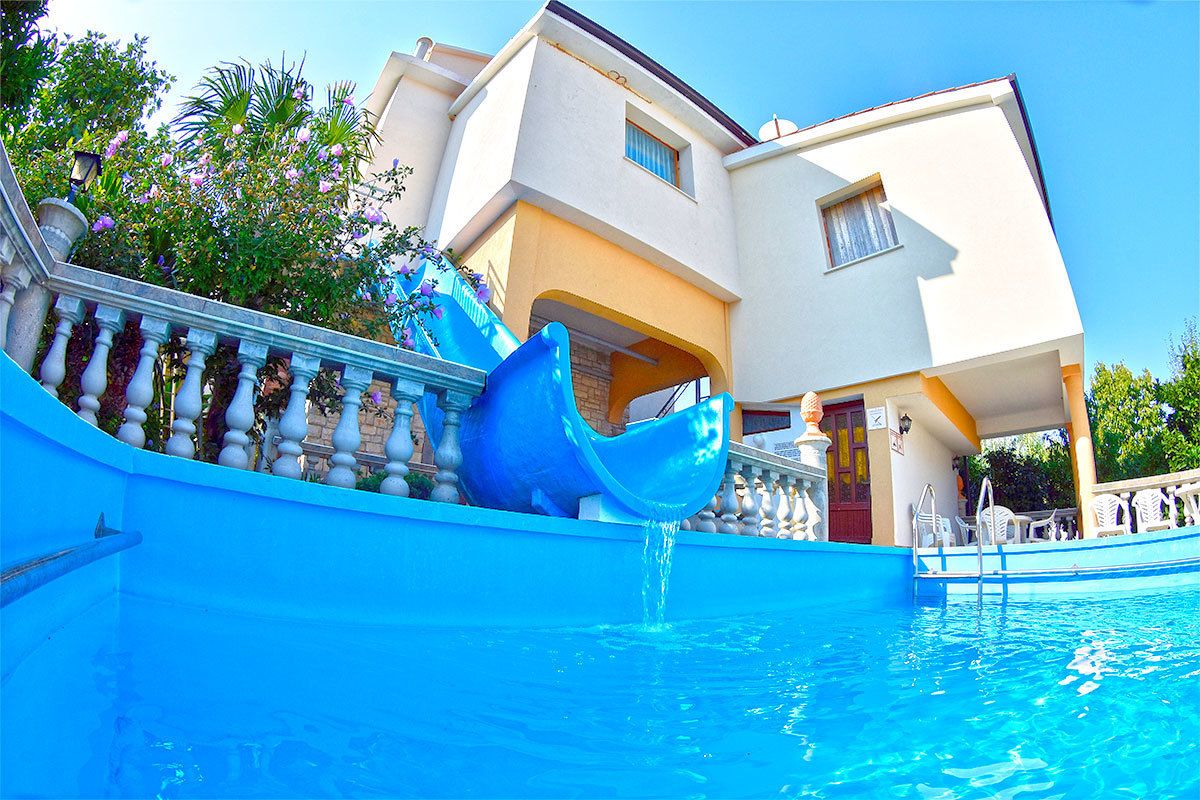 Una piscina con un tobogán azul, un edificio con balcón y un cielo despejado.