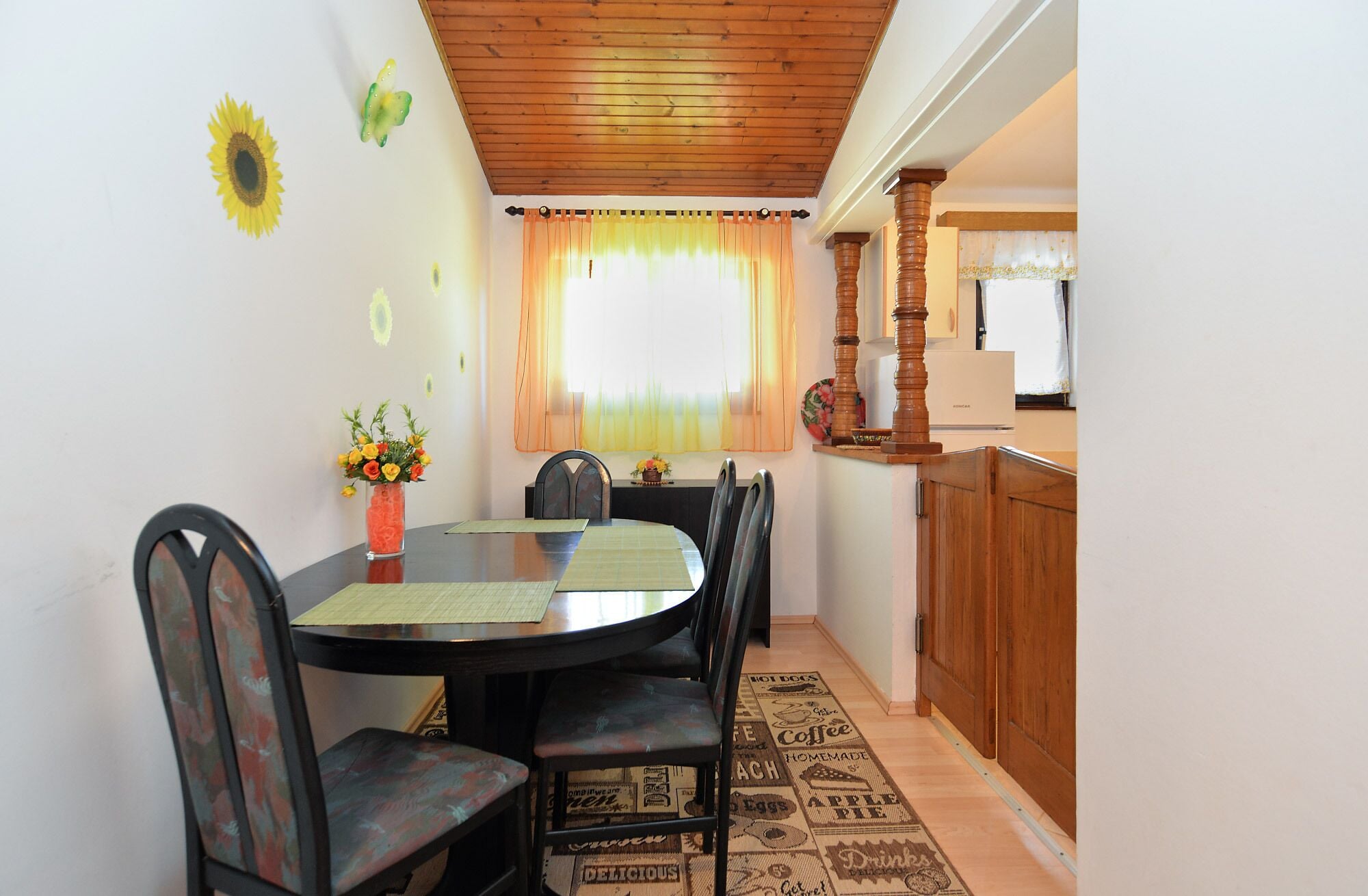 A dining area with a round table, chairs, a vase of flowers, and a wooden ceiling.