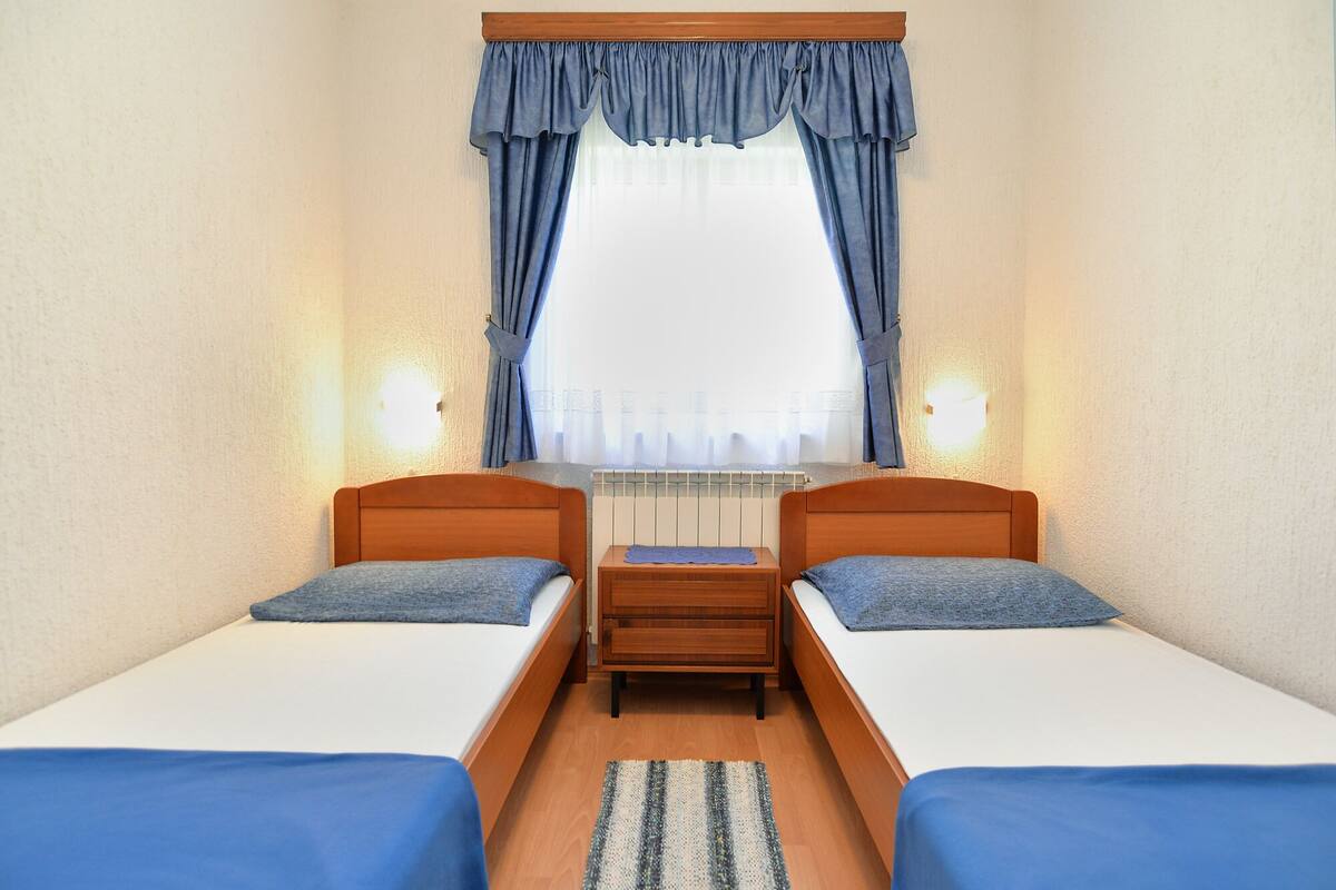 A room with two single beds, a small wooden nightstand, and a window with blue curtains.