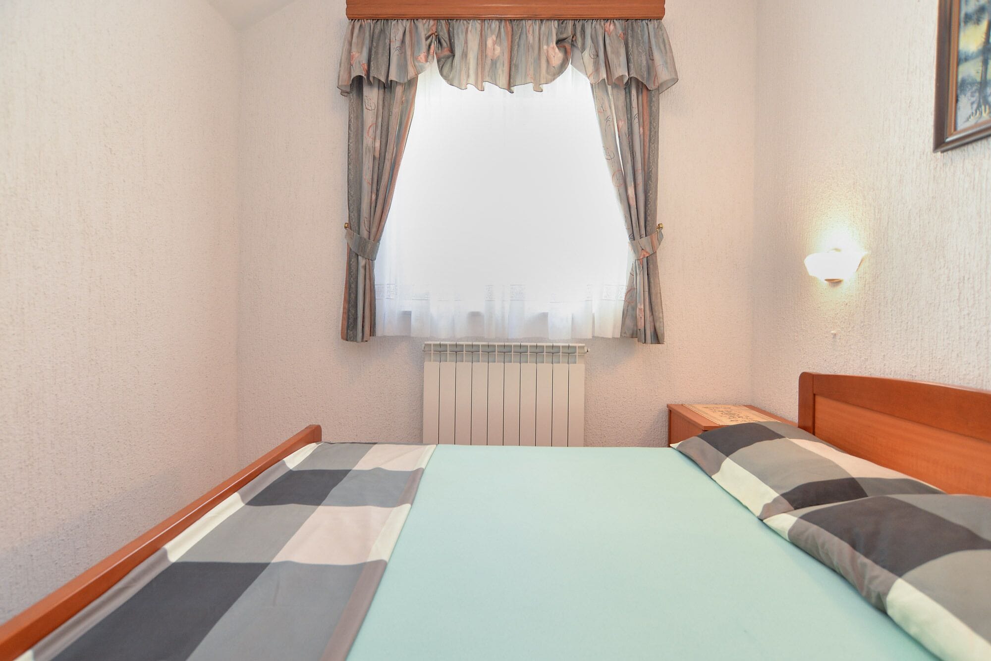 A bedroom with a bed, a window with curtains, a radiator, and a wall-mounted light.