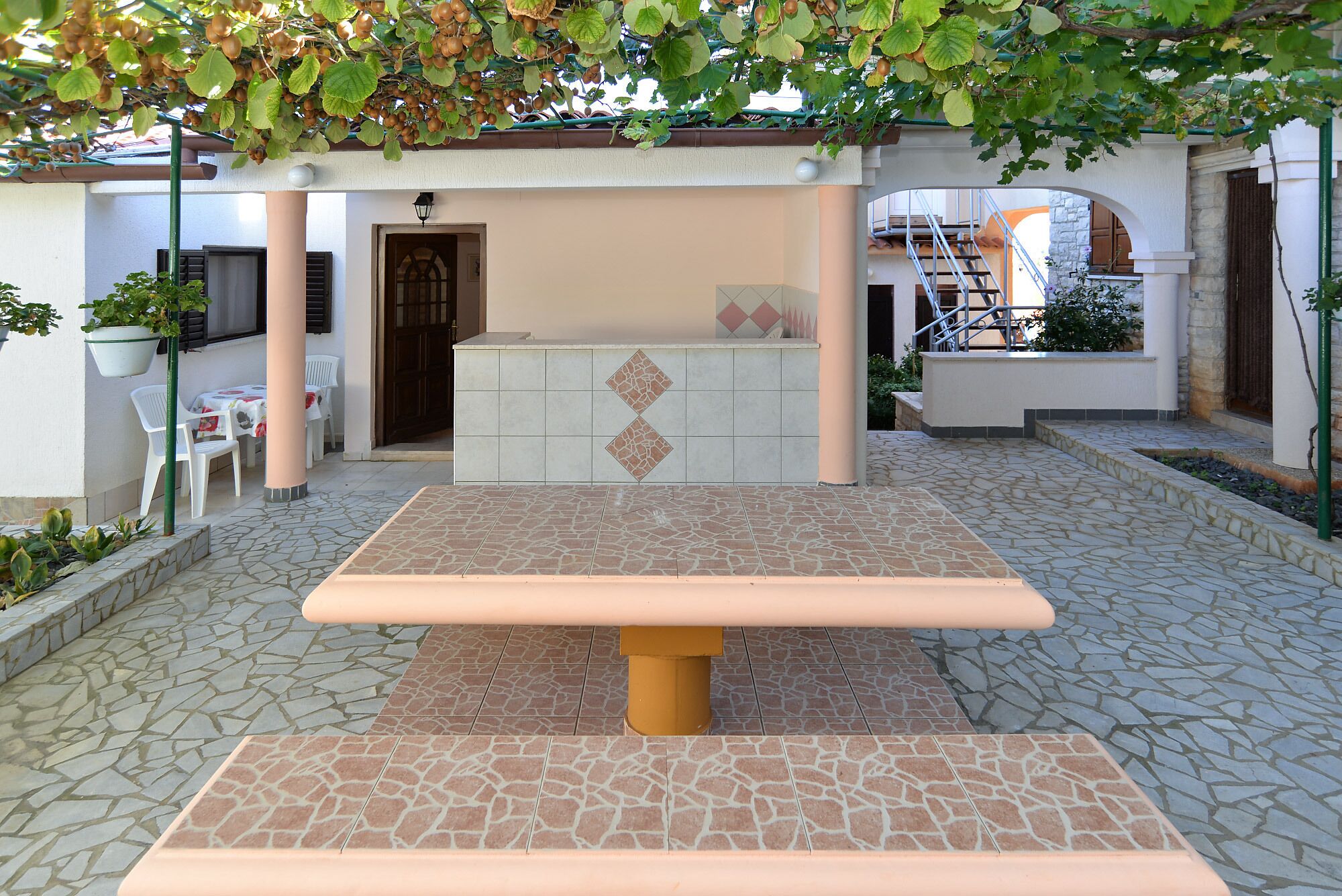 Eine Außenpatio-Terrasse mit Tisch und Stühlen, einem gefliesten Boden und einer Pergola mit hängenden Pflanzen.
