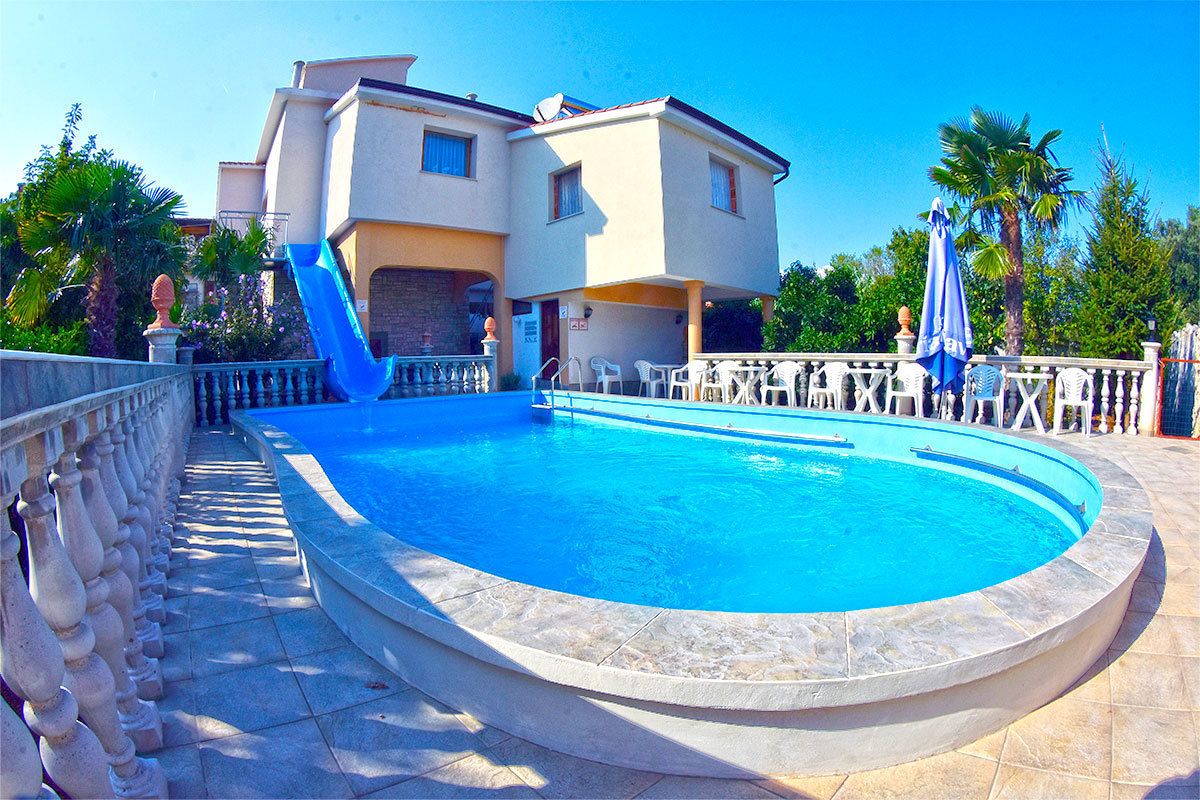 A swimming pool with a stone deck, a white building with a balcony, and palm trees in the background.