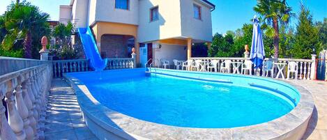 A swimming pool with a stone deck, a white building with a balcony, and palm trees in the background.