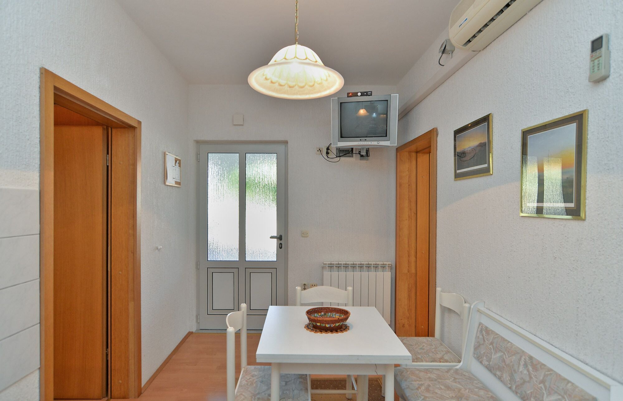 A small dining area with a white table, two chairs, a bowl on the table, a pendant light, a television on the wall, and a radiator below.