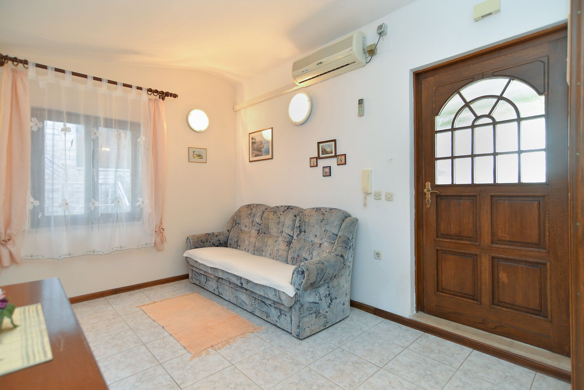 Una habitación con puerta de madera, ventana con cortina, sofá, alfombra pequeña y aire acondicionado de pared.