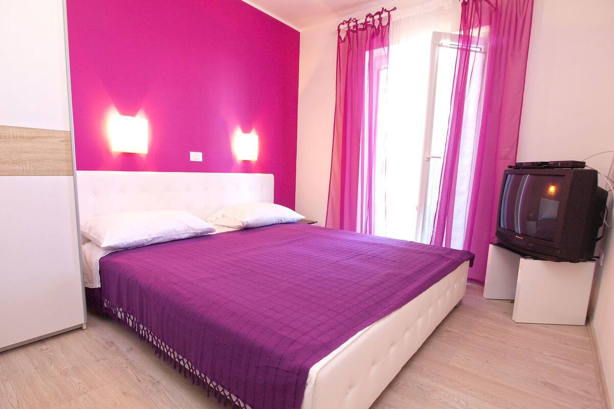 Un dormitorio con una cama grande, un televisor sobre un mueble y una pared de color rosa brillante.