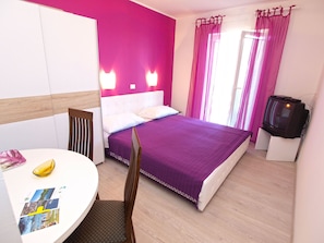 Quarto de hotel com uma cama, uma televisão, uma mesa com uma banana e folhetos, e uma cadeira.