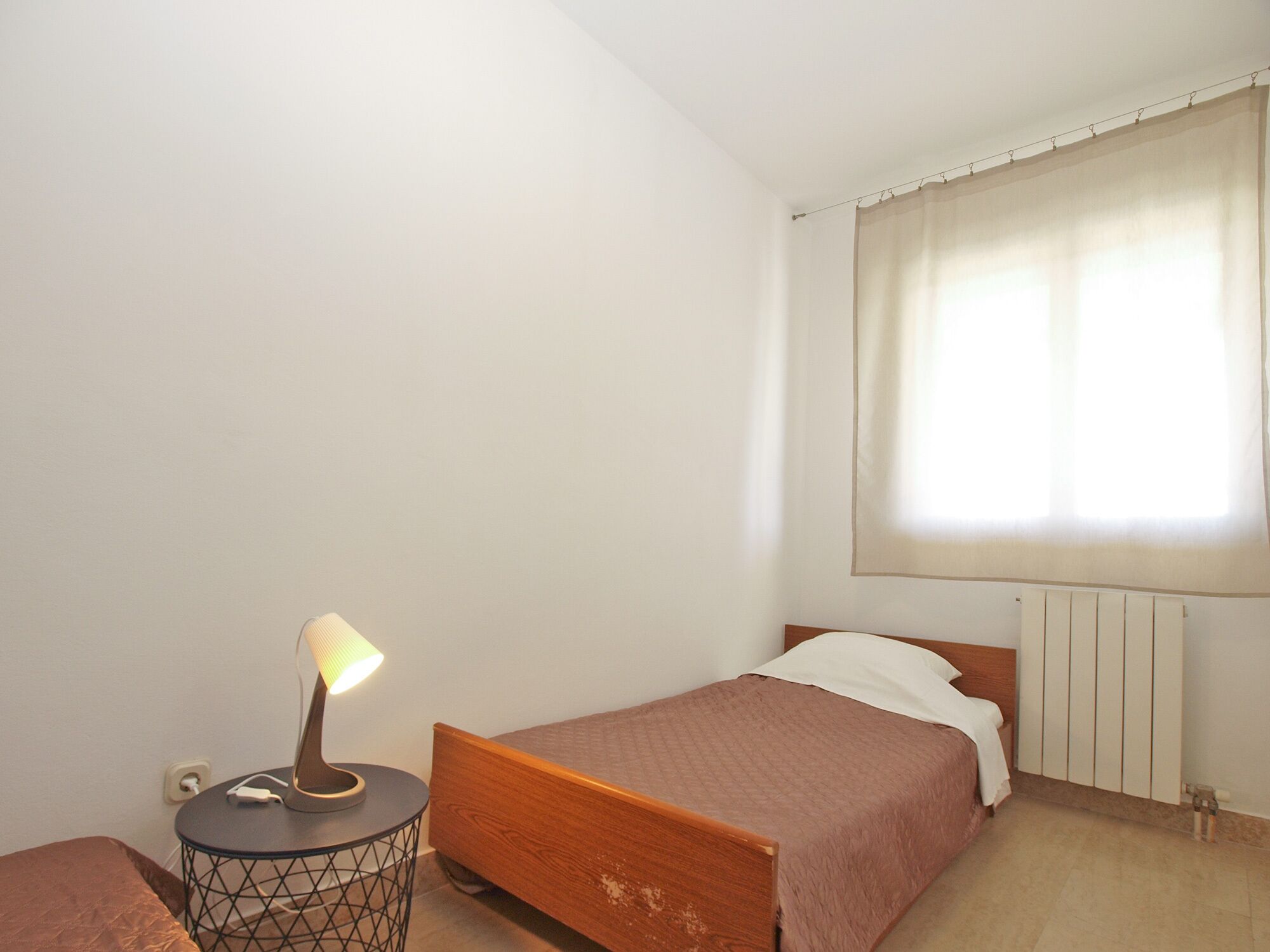 A bedroom with a single bed, a bedside table with a lamp, a radiator, and a window with curtains.