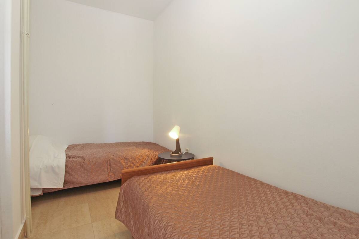 A small, dimly lit room with two single beds, a bedside table with a lamp, and plain white walls.
