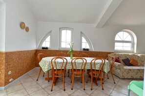 A dining area with a table set for six, surrounded by six chairs with a fruit pattern. There are two arched windows providing natural light, and a sofa with patterned cushions in the background. - Apartments Nelli 624 (Pula)