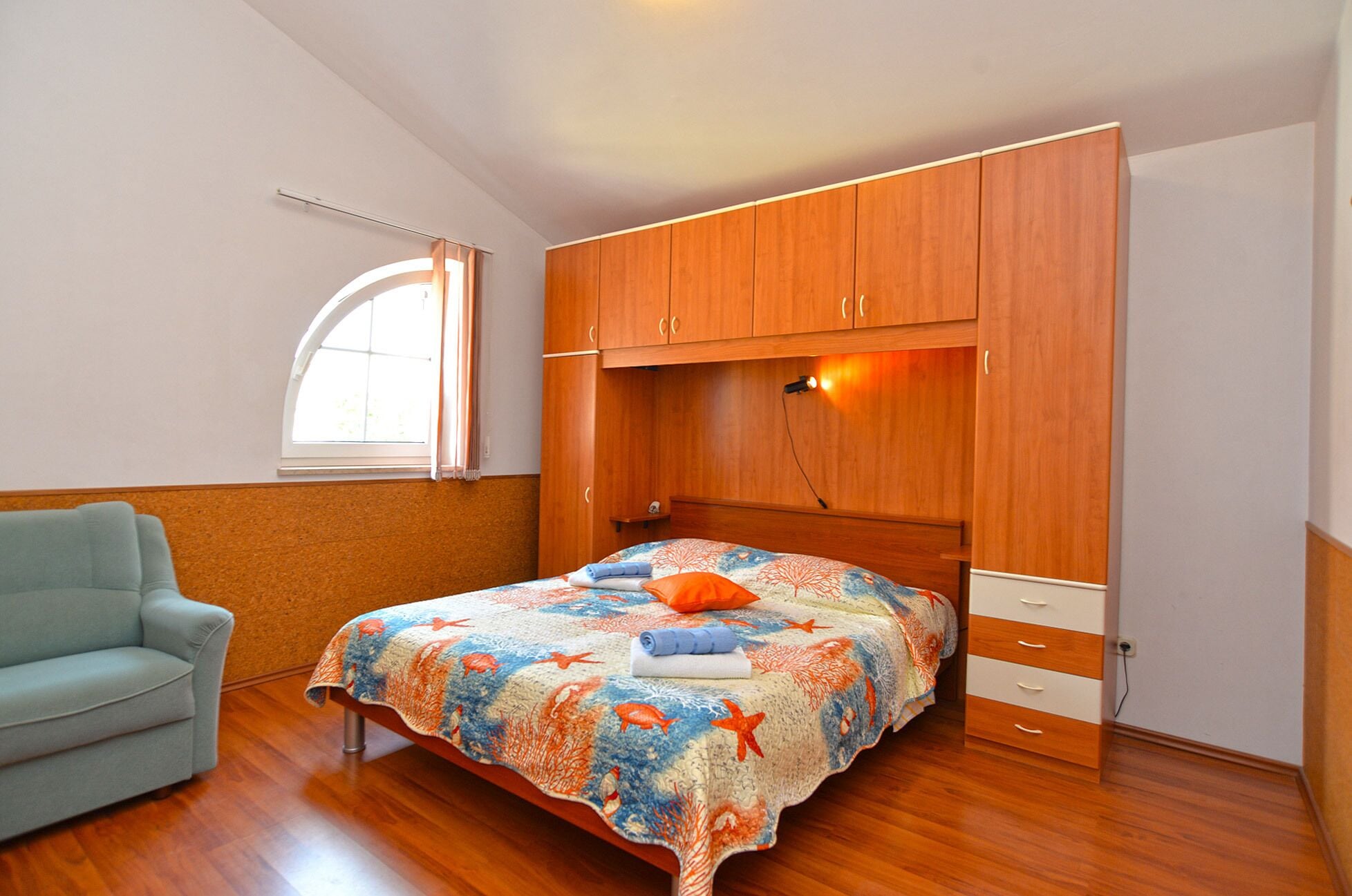 A bedroom with a bed, a light blue armchair, wooden wardrobe, and a window with curtains.