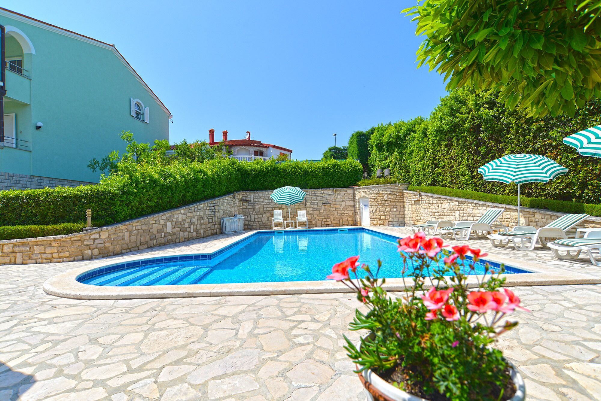 A swimming pool with stone tiles, surrounded by lounge chairs and umbrellas, with a stone wall and greenery in the background.