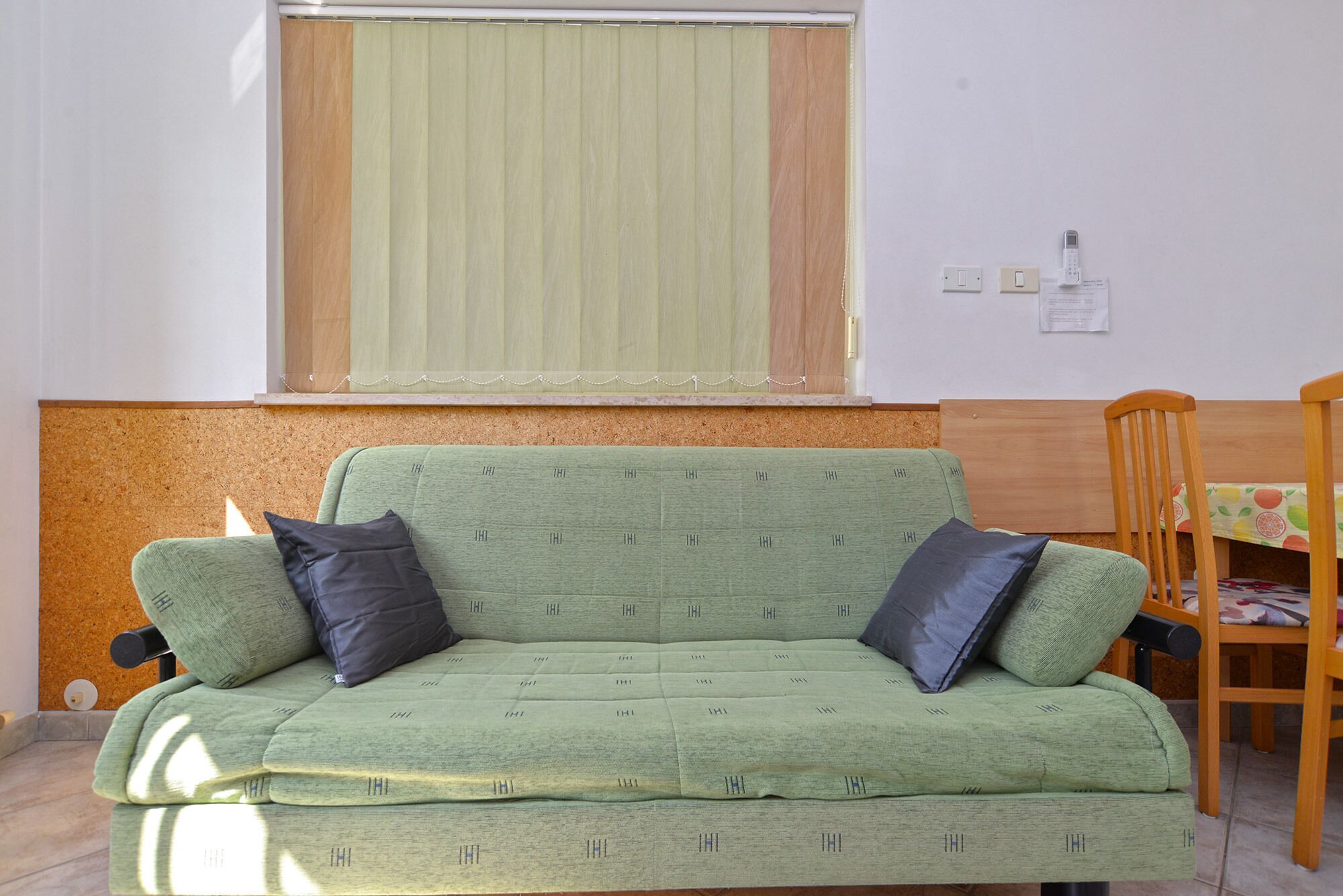 A green sofa with two black cushions, a wooden chair, and a cork bulletin board.