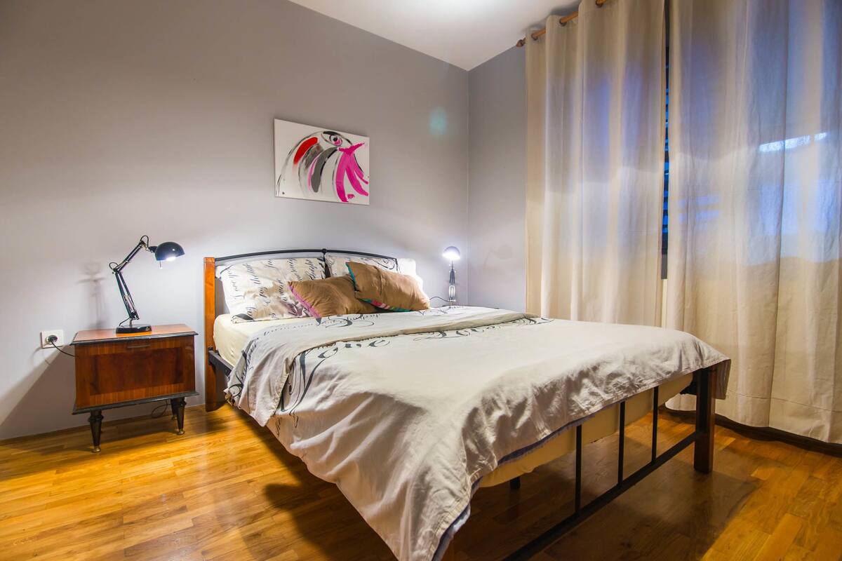 Un dormitorio con cama, mesita de noche, lámpara y una obra de arte en la pared.