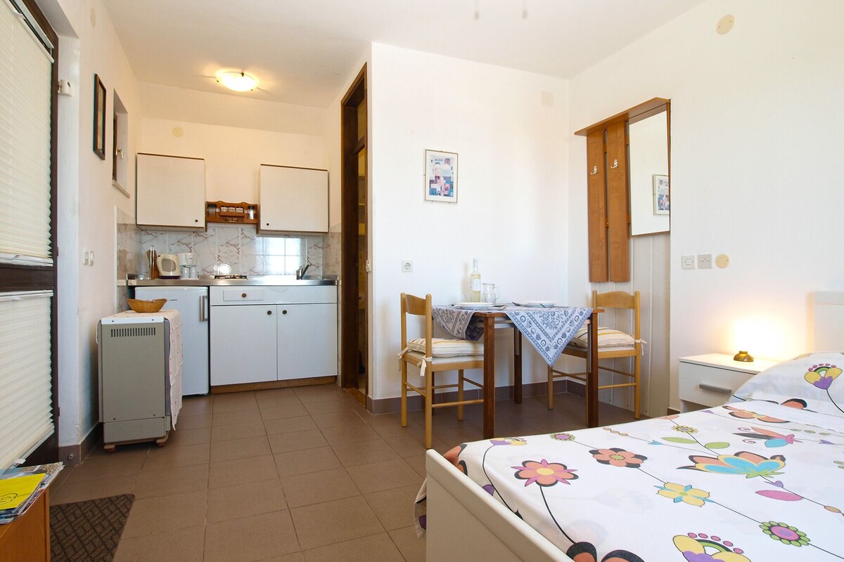 A compact, well-lit room with a kitchenette, a dining area with a table and chairs, and a bed with a floral bedspread.