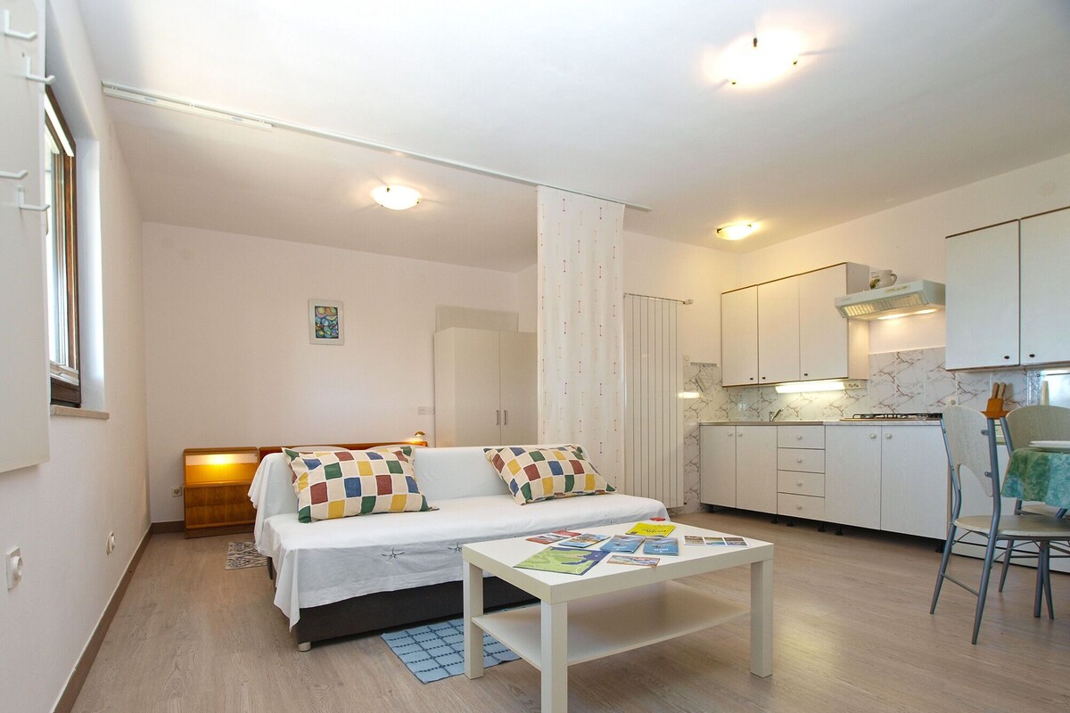 A compact living space with a bed, a small table, a kitchenette, and a window.
