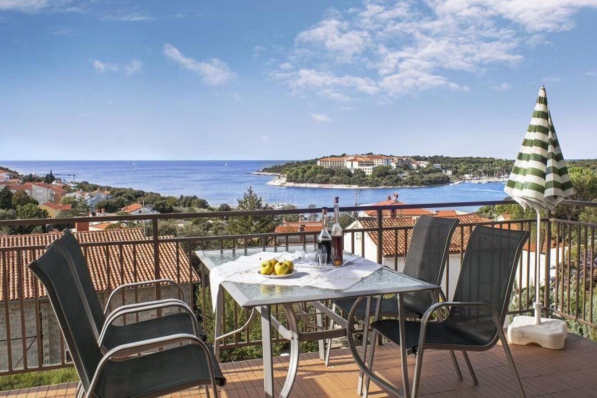 A balcony with a table set for two, overlooking a coastal view with a building and boats.