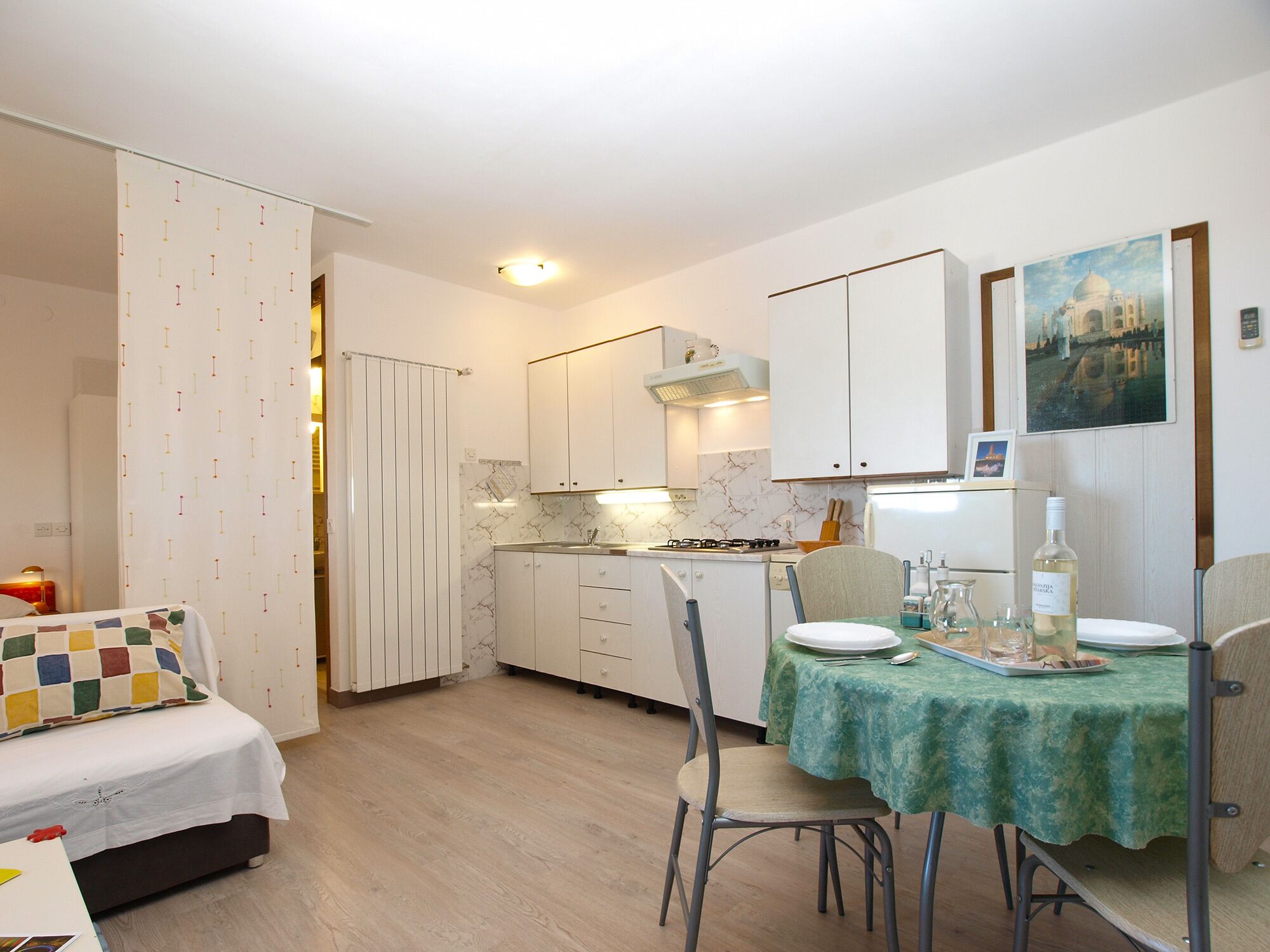 A compact kitchen with white cabinets, a dining table with a green tablecloth, and a bed with a colorful quilt.