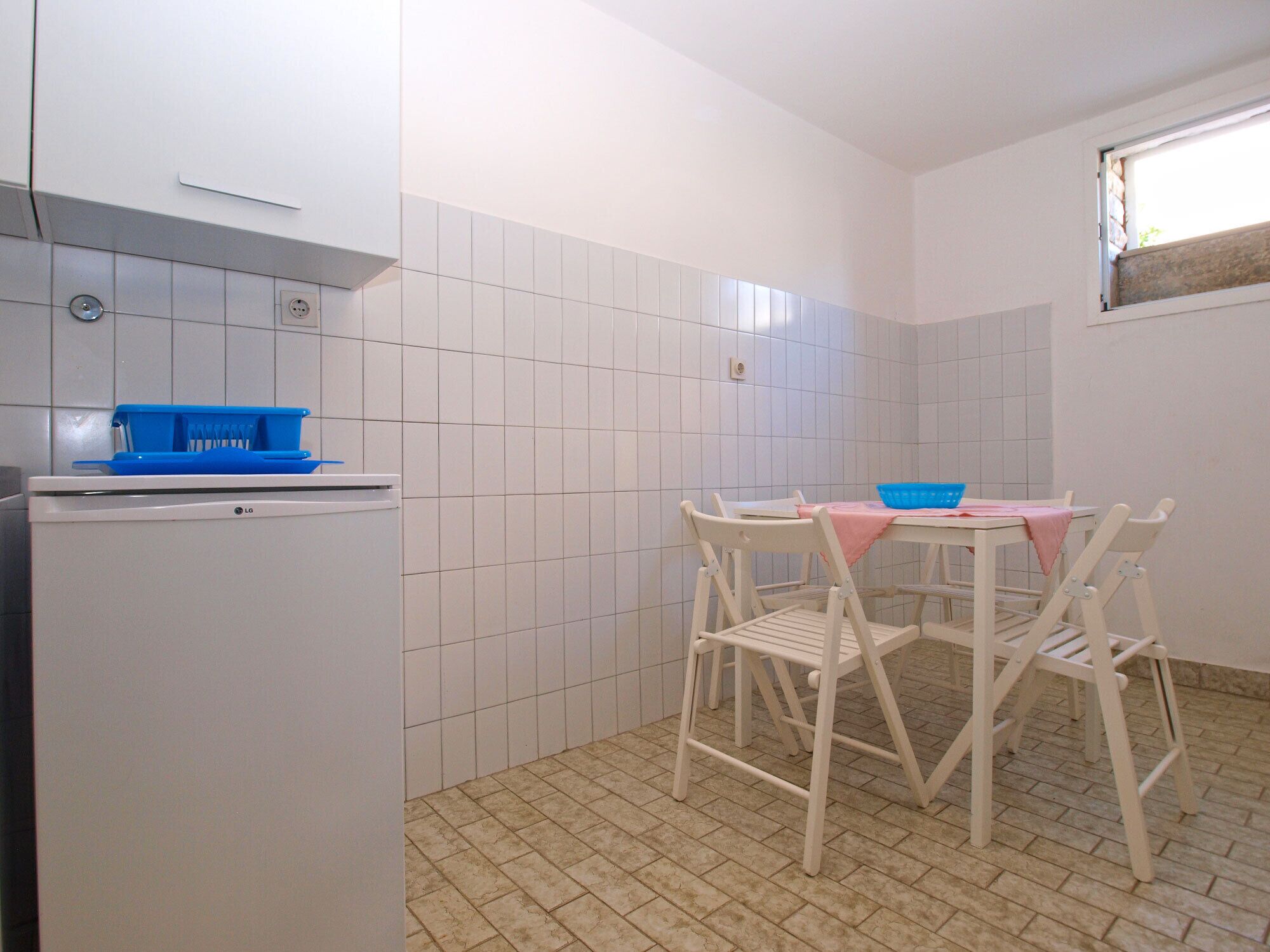 A small kitchen with white tiled walls, a white refrigerator, a table with chairs, and a window with a view.