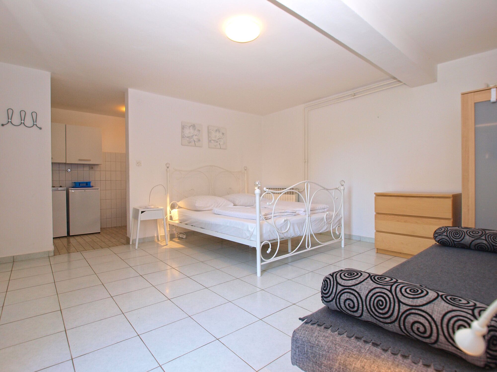 Een slaapkamer met een bed, nachtkastje, dressoir en een kitchenette op de achtergrond.
