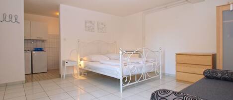 Een slaapkamer met een bed, nachtkastje, dressoir en een kitchenette op de achtergrond.