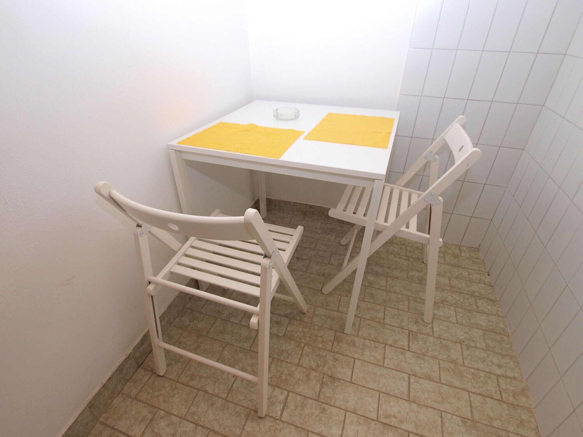 A small white table with two yellow placemats and two white chairs in a corner of a room with tiled flooring.
