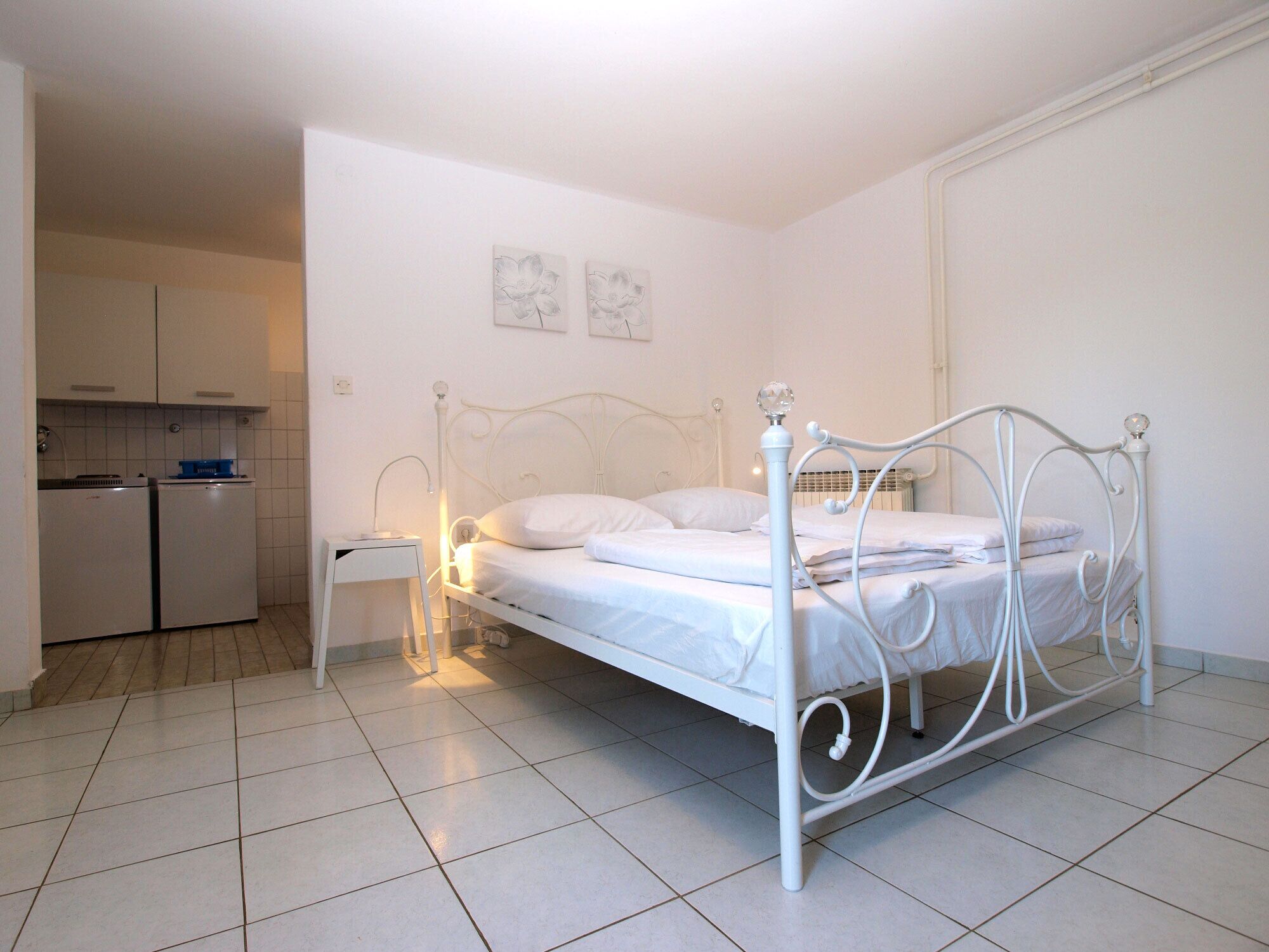 Una cama con dosel de metal blanco, un mesita de noche con lámpara, una pequeña cocina americana con nevera y hornilla, y piso de baldosas.