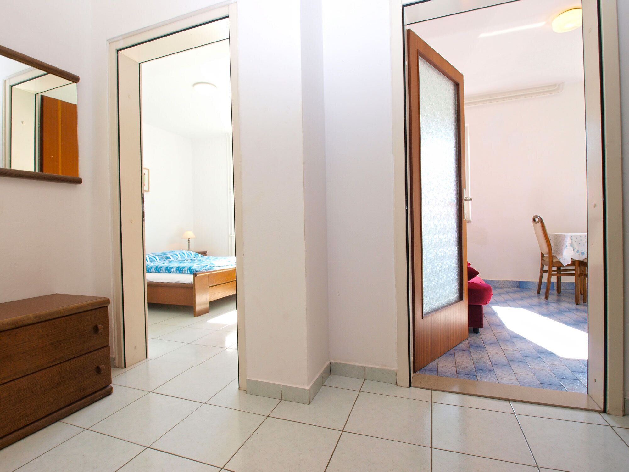 A room with a bed, a wooden dresser, and a door leading to another room with a table and chairs.