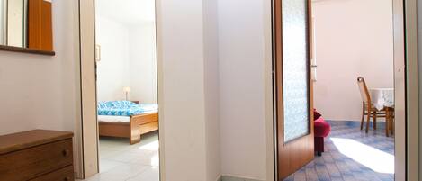 A room with a bed, a wooden dresser, and a door leading to another room with a table and chairs.