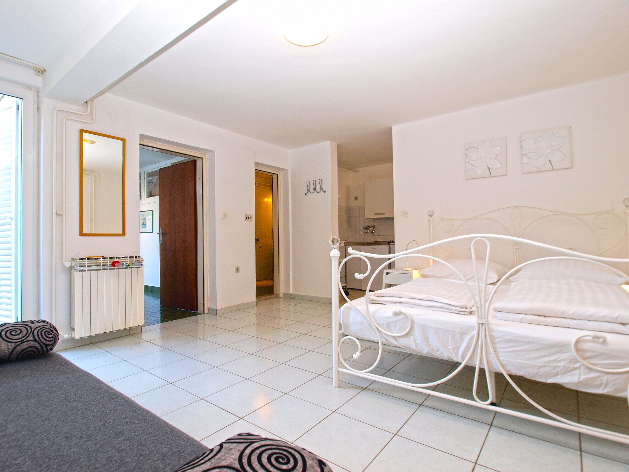 Een slaapkamer met een bed van wit metaal, een grijze bank, een radiator en een deur naar een badkamer.