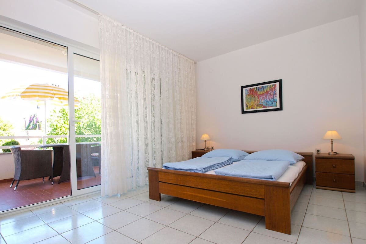 A bedroom with a bed, two bedside tables with lamps, a window with curtains, and a painting on the wall.