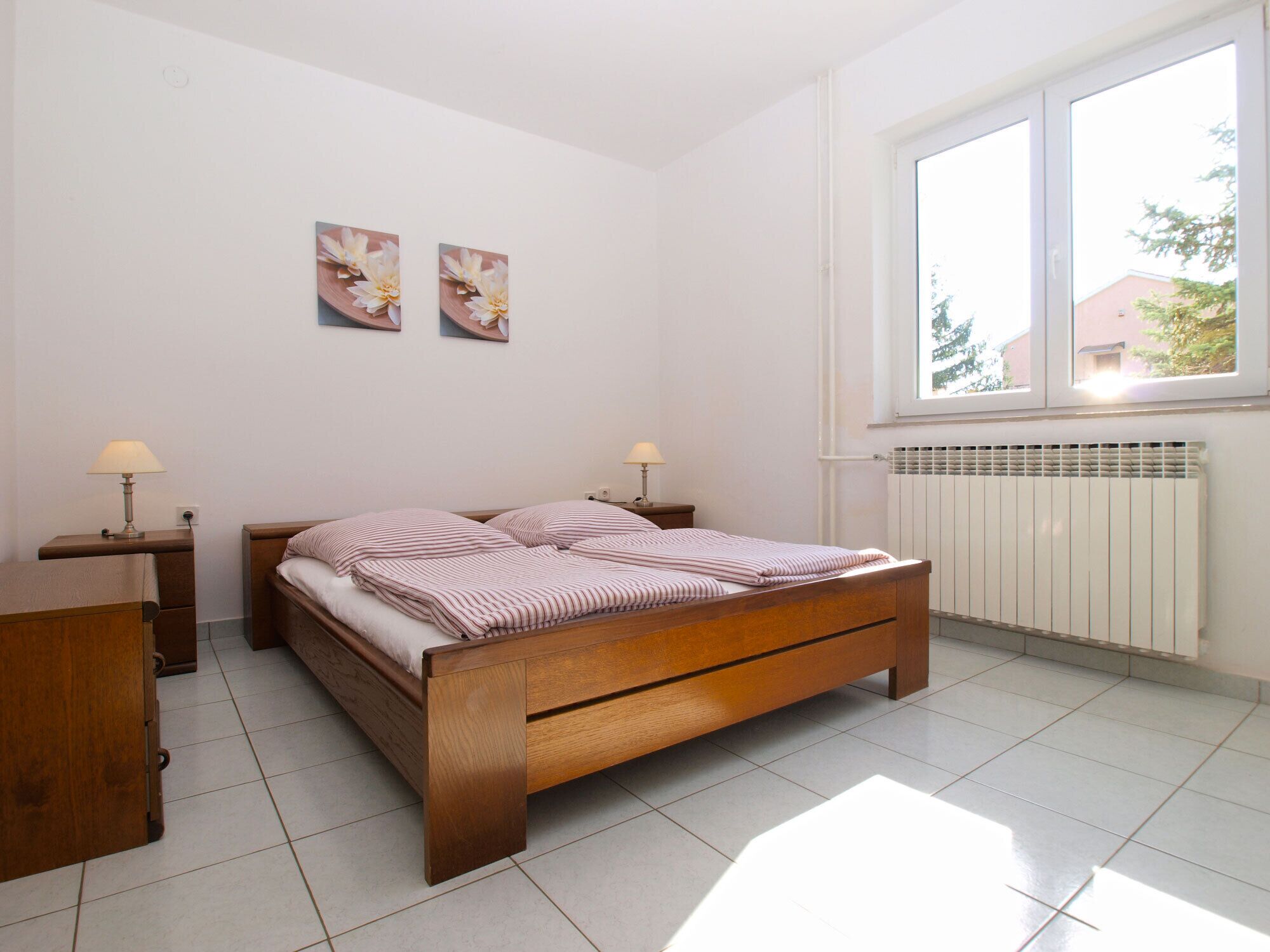 A bedroom with a wooden bed, two bedside tables with lamps, a radiator, and a window with a view of buildings.