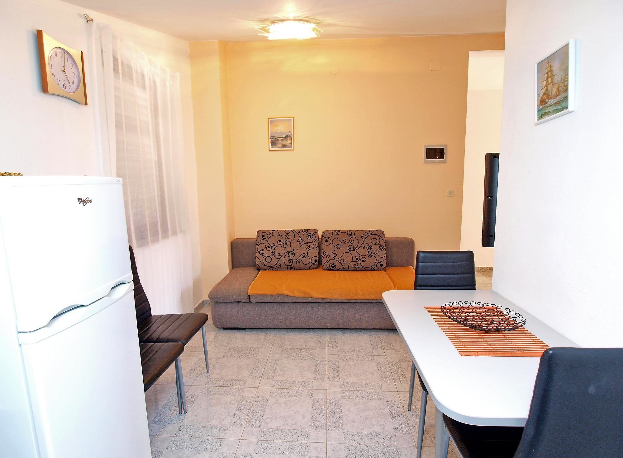 A compact living space with a kitchenette, a sofa with patterned cushions, a dining table with a decorative item, and a wall-mounted clock.