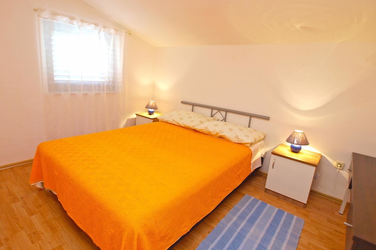 A bedroom with a bed, nightstands, a lamp, a window with curtains, and a wooden floor.